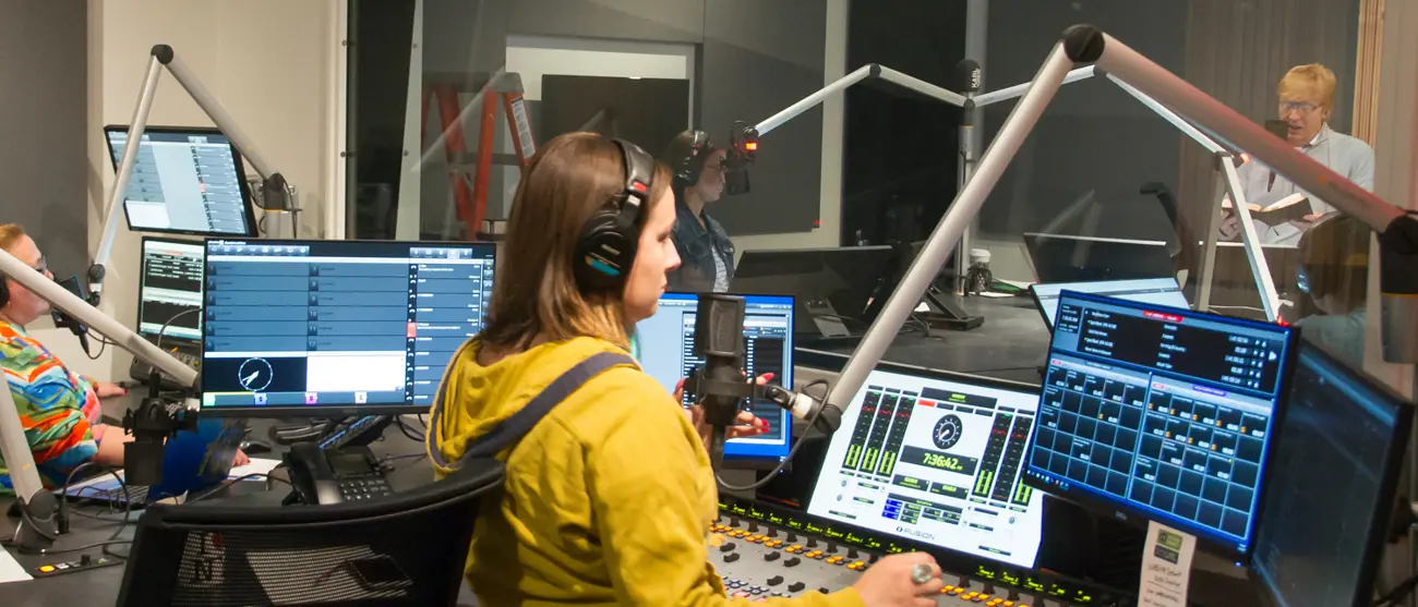 Four people work in a radio broadcasting room as one man reads from a book into his microphone.  